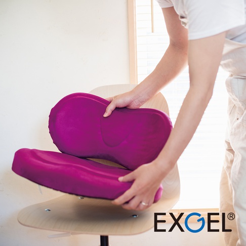 雙手正在放置一個紫色的 HUG COMFY PREMIUM 背部支撐坐墊，坐墊為分層設計，材質看起來柔軟有彈性，整體呈現符合人體工學的曲線。