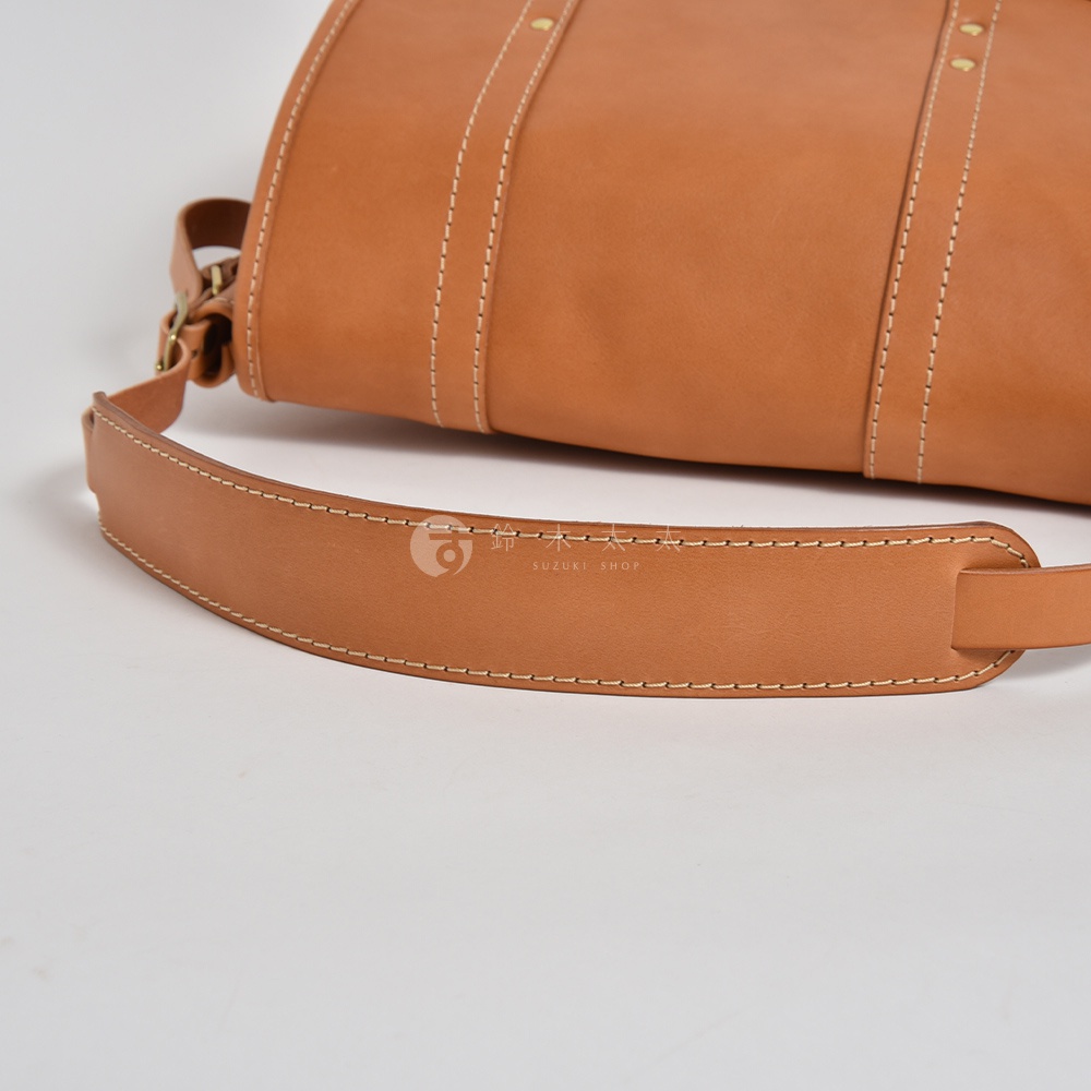 職人手作皮革復古郵差包，採用天然皮革製成，呈現溫潤的焦糖棕色。包身設有雙條加固皮帶，以白色縫線裝飾，搭配可調節的寬版皮革肩帶，展現經典復古風格。