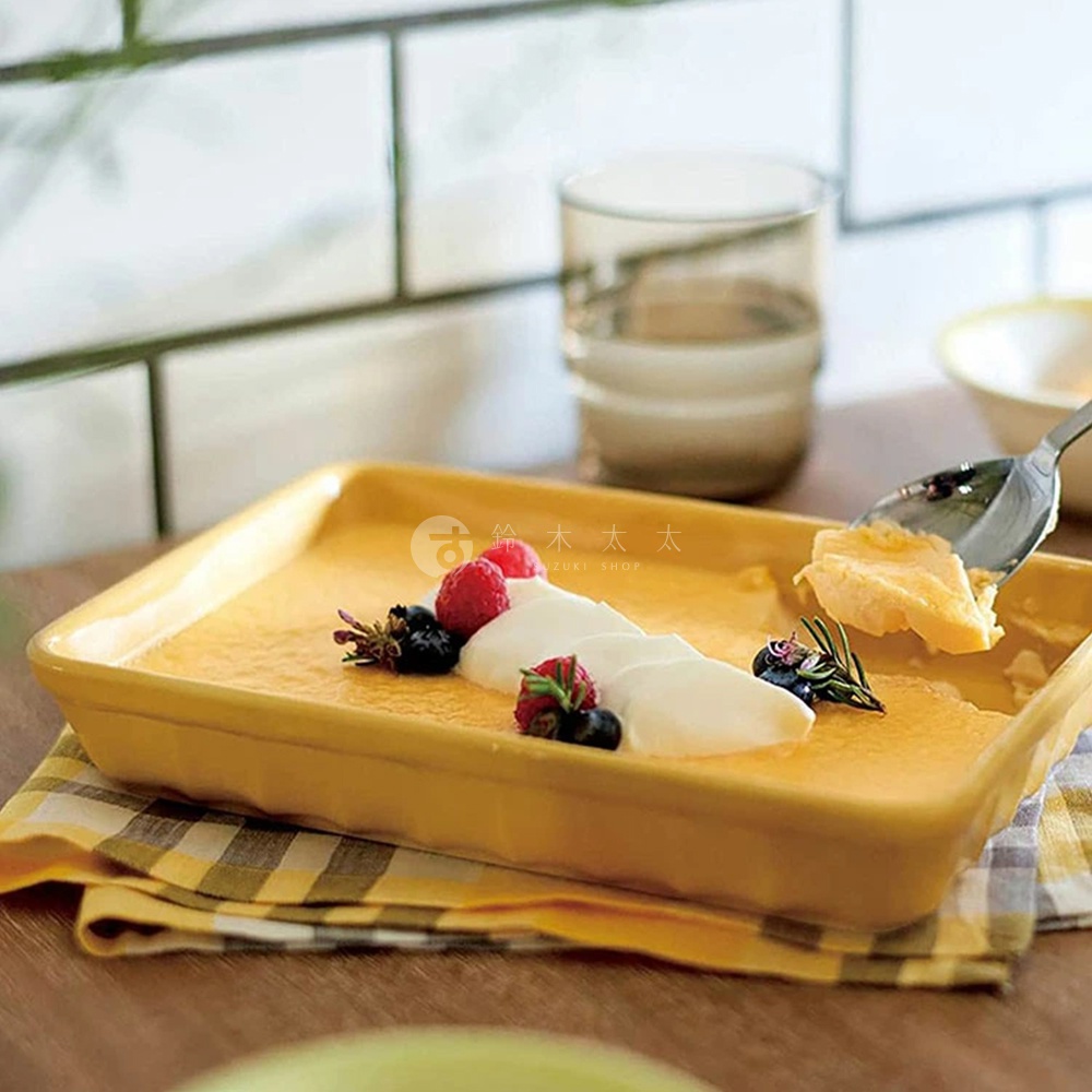黃色美濃燒釉彩萬用長方烤盤，內盛布蕾，表面裝飾有奶油、覆盆子、藍莓與迷迭香，烤盤置於格紋餐巾上。
