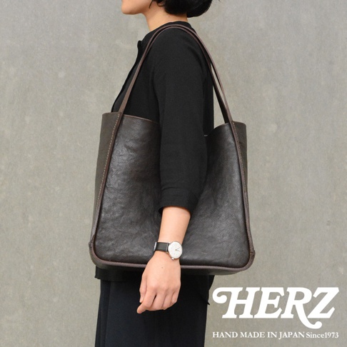 一位女性側身展示日本HERZ職人手作的深棕色皮革托特包，該包款呈現簡約的方形立體結構與自然皮革紋理，搭配纖細的肩背帶，整體風格低調沉穩，右下角印有HERZ HAND MADE IN JAPAN Since1973字樣。