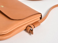 職人手作皮革橫式隨行袋，淺棕色，可見其縫線、翻蓋和金屬釦。袋子由光滑的皮革製成，附有可調節的皮革背帶，透過皮革繩結固定。