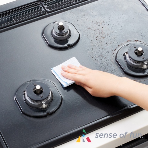 手持白色帶有藍色邊緣的清潔巾，正在擦拭黑色瓦斯爐表面上的污漬。