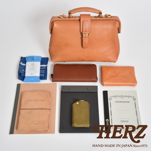 一組職人手作皮革配件，包含一個大型的駝色手提包，側邊有金屬扣環，包身配有皮帶和金屬扣。另有兩個長夾，一個為深棕色，一個為淺棕色。配件包含藍色印花布、藍色濕紙巾包、粉紅色筆記本、黑色文件夾、綠色鑰匙包以及一本淺灰色的筆記本。