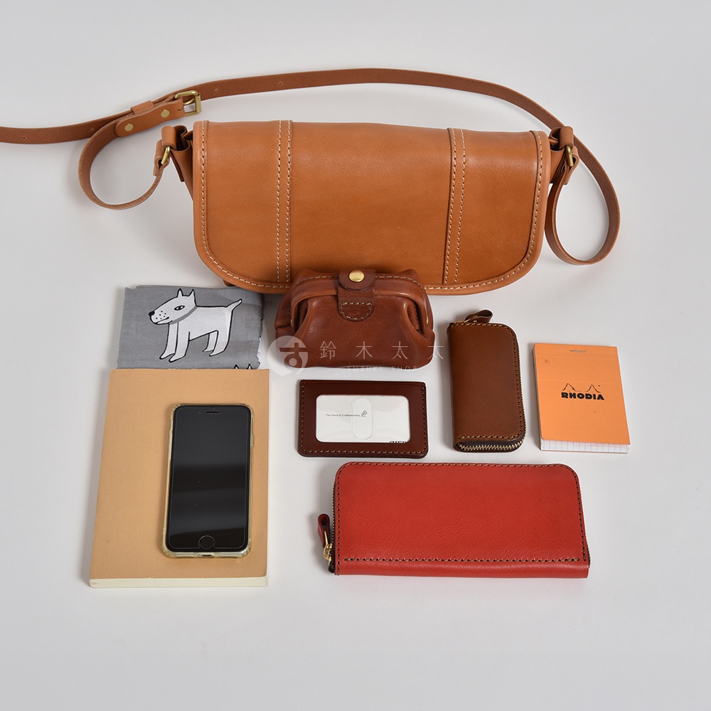 職人手作皮革復古郵差包，以焦糖色光滑皮革製成，縫線清晰。包內展示多樣配件，包括棕色皮革零錢包、卡夾、鑰匙包、紅色長夾，以及印有狗狗圖案的灰色布巾，還有一個淺棕色筆記本和手機。