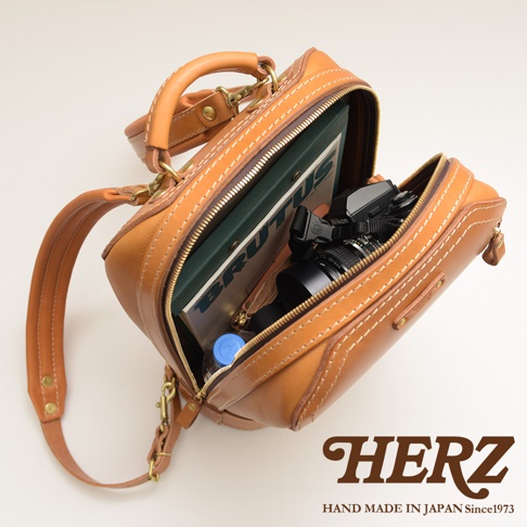 焦糖色皮革製成的立體商務後背包，主隔層打開，露出雜誌、水瓶和相機。包款採用手縫白色縫線，配有頂部提把和可拆卸肩帶。