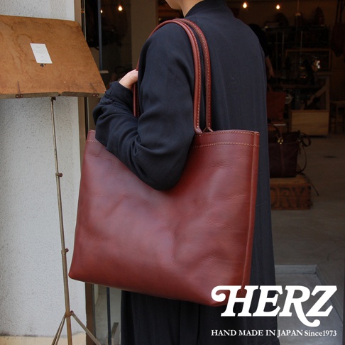 一位身穿黑色長袍的人士，肩上斜背一個深棕色皮革托特包，包包的縫線清晰可見，包身線條簡潔大方，整體呈現出職人手作的質感。