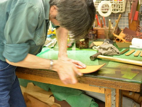 職人正在工作室的綠色工作檯上，用工具打磨一件淺棕色的皮革物品，旁邊散落著各種皮革材料、工具和配件。