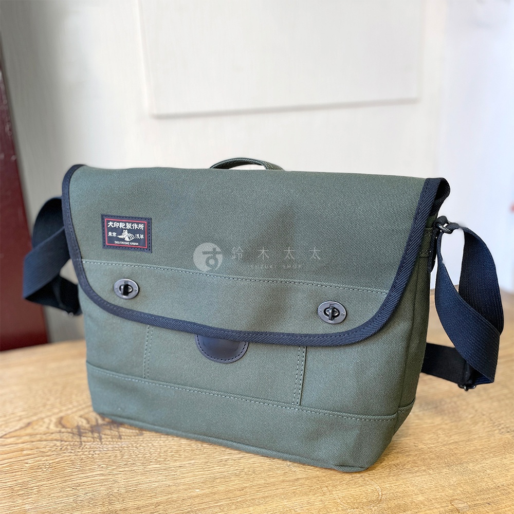 綠色純棉帆布雙扣郵差包，配有黑色邊緣滾邊和黑色肩帶。包蓋上有一個印有日文的方形標籤。