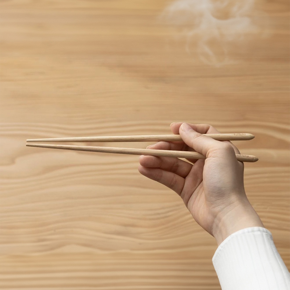 一雙 23 公分長的楓木圓筷，由右手握持，筷子末端有品牌標誌，背景為淺木紋桌面。