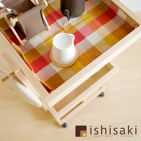 淺色木製收納推車，頂層鋪著紅、黃、橘格紋布，擺放一個白色馬克杯、木製杯墊及一個深色水壺。推車側面掛有白色布袋。