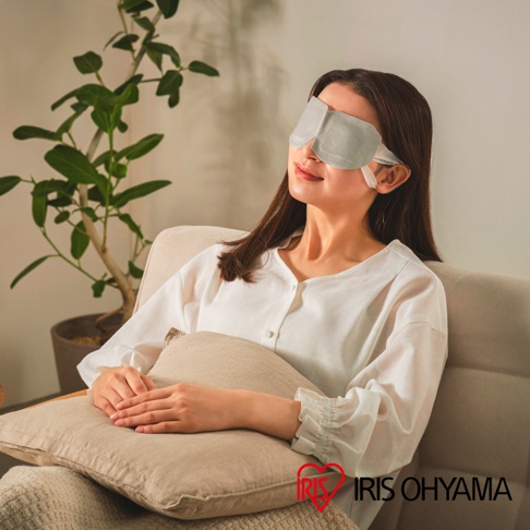 一位年輕女性坐在米色沙發上，頭戴淺灰色IRIS OHYAMA蒸氣眼罩，閉目養神並抱著一個米色抱枕，旁邊擺放著一盆室內盆栽，畫面呈現出舒適、放鬆的生活氛圍。