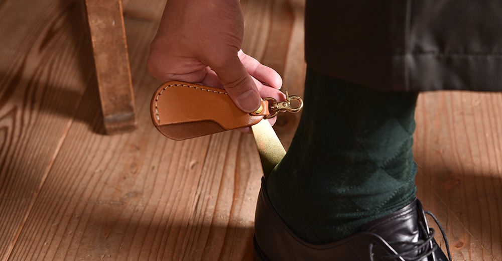 職人手作皮革隨身金屬鞋拔，淺棕色皮革搭配黃銅掛扣，金屬鞋拔部分為金色，使用者正將鞋拔插入黑色皮鞋。