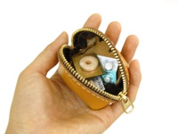 手持一只 tan 色皮革貝殼造型零錢包，拉鍊敞開，內部可見藥丸藥片，零錢包由皮革製成，內襯為深棕色。