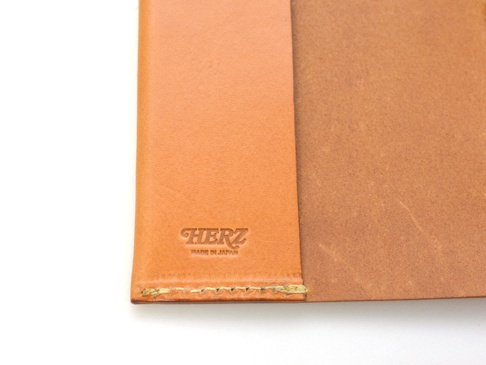 職人手作皮革附繩書套，淺棕色天然皮革，帶有壓印的「HERZ」品牌標誌及「MADE IN JAPAN」字樣，邊緣縫線工整。