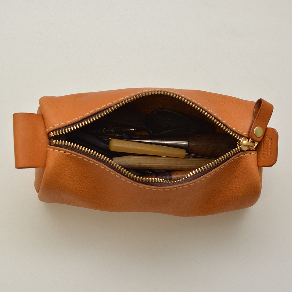 職人手作皮革多用途收納筆袋，採天然皮革材質，顏色為焦糖棕。袋口拉鍊呈金黃色，內有筆、尺、裁刀等文具。