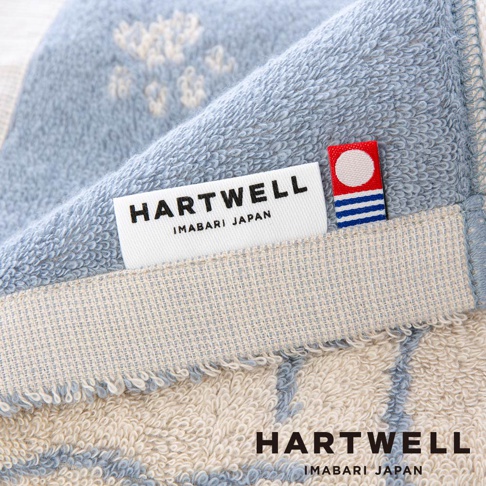 這是一張 Hartwell 今治午睡貓咪雙色毛巾的特寫鏡頭，照片中毛巾呈現藍色與米白色交織的質感，布面上飾有淺色的貓咪圖案刺繡，邊緣處縫有精緻的 Hartwell 品牌標籤，標籤旁飾有標誌性的紅圓與藍色線條小標，展現出高品質日式織物的細節與柔軟手感。