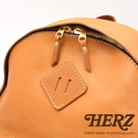 職人手作皮革後背包，近拍展示其焦糖棕色皮革材質、雙拉鍊設計以及方形皮革飾片，搭配精緻的縫線細節。