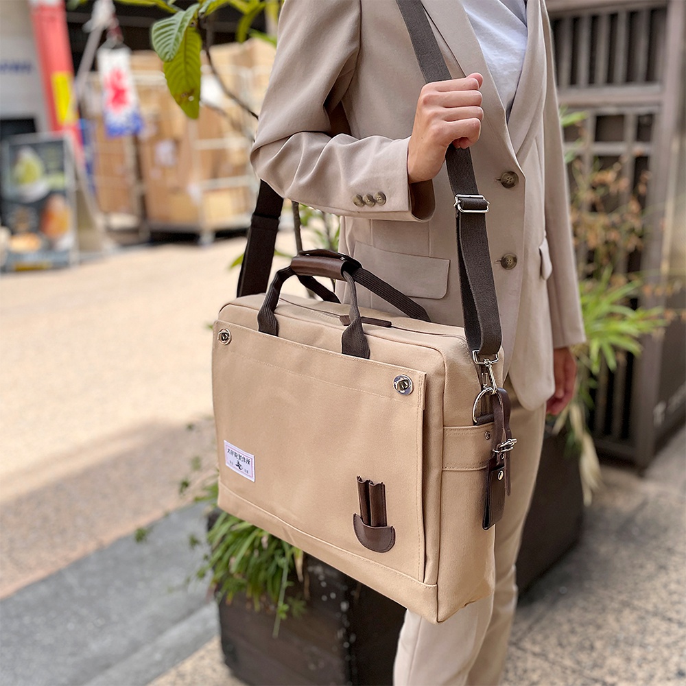 米色純棉帆布商務公事包，B4尺寸，配有棕色皮革提把與可拆卸肩帶，正面有兩個鈕扣式翻蓋口袋及一個皮革筆夾。