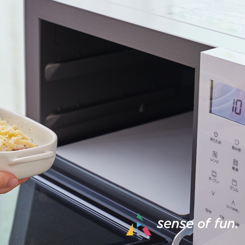 一隻手正將裝有食物的白色烤盤放入微波爐的爐腔內，微波爐面板上顯示時間和多個烹飪功能按鈕。