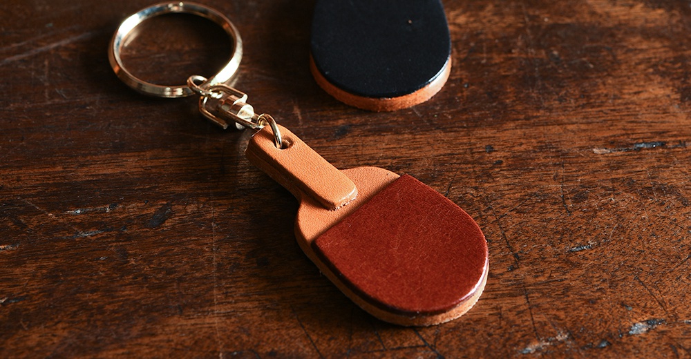 職人手作皮革桌球拍造型鑰匙圈，以棕色皮革製成，搭配金色金屬掛環與鉤扣，造型小巧精緻，可掛於包包或鑰匙上。