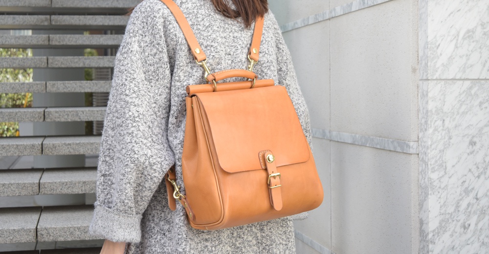 展示一個淺棕色皮革後背包，採用屋瓦造型設計，配有金色金屬扣環與調節式肩帶。