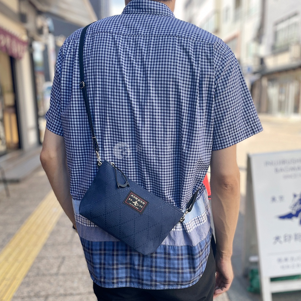 深藍色刺子織斜背包，有菱形縫線紋路，正面有一個標籤，斜背包以黑色細肩帶斜跨在穿著藍白格紋襯衫的男子身上。