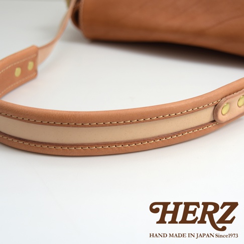 一條 tan 色皮革郵差包的肩帶，帶有雙色拼接和縫線細節，以及兩個黃銅扣環。