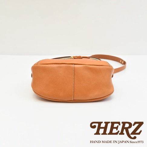 HERZ 品牌標誌，下方標有 HAND MADE IN JAPAN Since1973，圖片中央是一個棕褐色皮革側背包的特寫，從下方視角拍攝，可見其圓潤的底部和縫線細節。