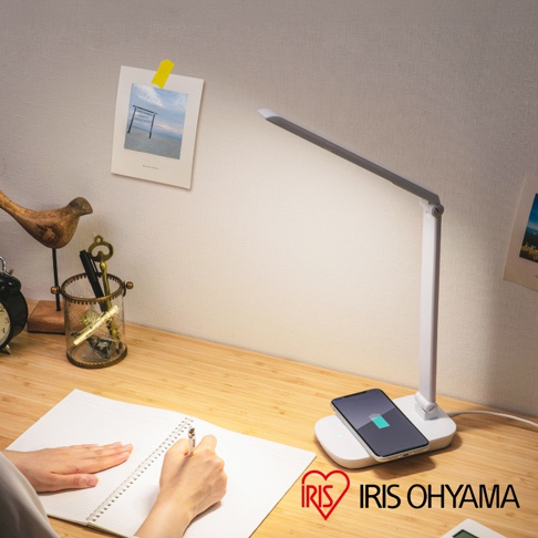 白色LED檯燈，燈臂可調角度，底部設有圓形無線充電盤，盤上放置一支手機，顯示充電中。檯燈與充電盤皆為簡約設計，材質為塑膠。