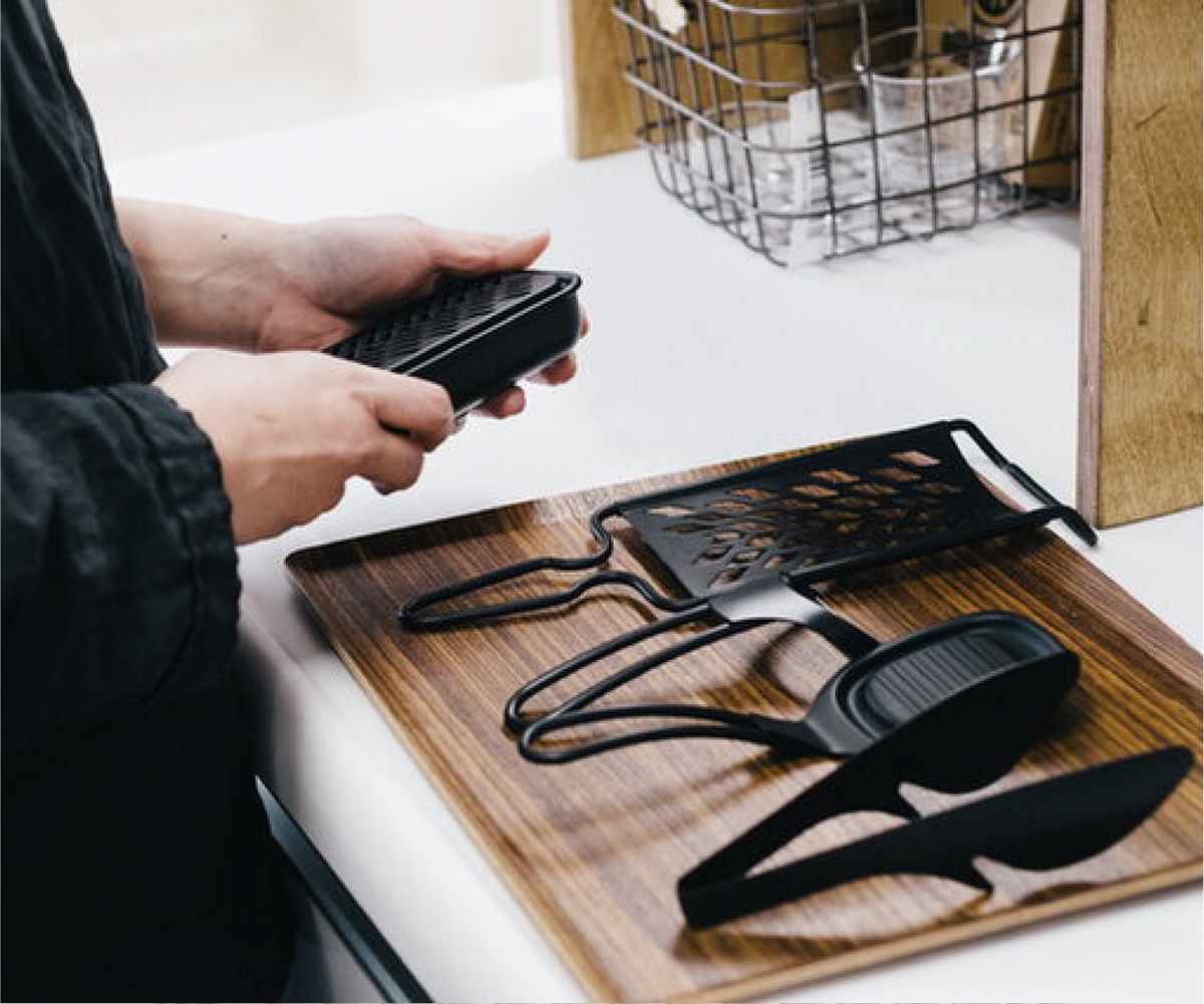 黑色安全刨絲器，包含一個刨絲網板和一個可收納刨絲網板的容器，以及一雙黑色夾子，所有物品放置在木質托盤上。