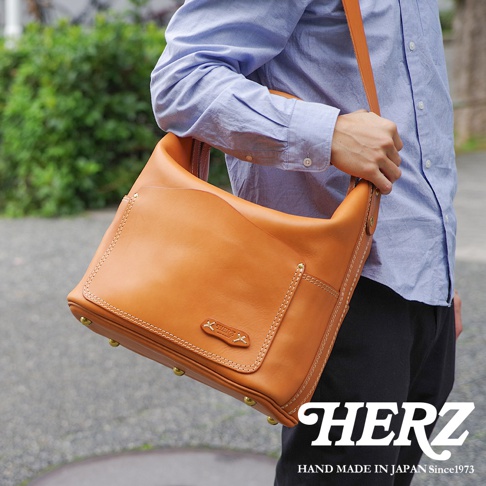 身著淺藍色襯衫的男子，肩揹一個棕色厚實皮革單肩波士頓包，包款有明顯的車縫線與金屬腳釘。