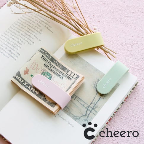 三款不同顏色 (粉色、淺綠色、淺黃色) 的「cheero」品牌萬能QQ變形夾，其中粉色夾子夾著一疊美金鈔票，淺綠色和淺黃色夾子則夾著乾燥植物，擺放在打開的書本上。