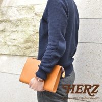 職人手作皮革三角底拉鍊手拿包，採用天然皮革材質，呈現自然光澤。包款為簡潔的長方形設計，配有拉鍊開合。手拿包的顏色為溫暖的棕褐色，與模特兒身上深藍色毛衣和灰色長褲形成對比。