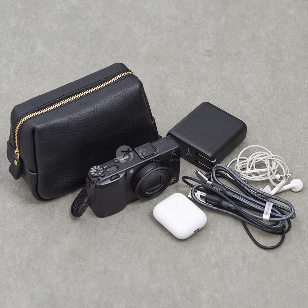 一張平鋪在灰色背景上的日本 HERZ 職人手作皮革收納包照片，旁邊擺放了一台黑色數位相機、黑色方型充電器、白色無線耳機充電盒、白色有線耳機以及黑色編織充電線。