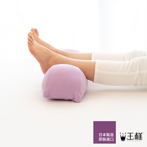 一對女性的雙腳抬高放在淺紫色、布質的半圓形抬腿枕上，腳踝和部分小腿被枕頭支撐。