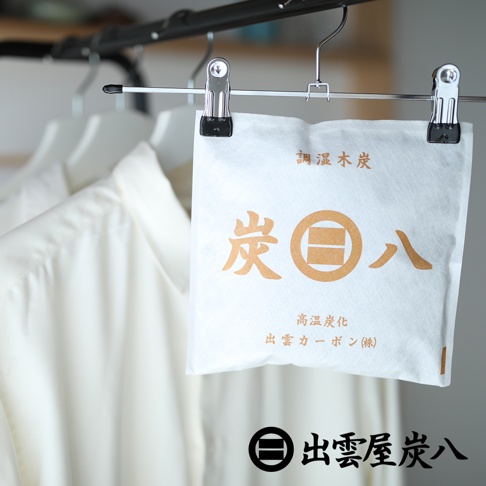 白色方形出雲屋炭八小型調濕木炭袋懸掛在衣架夾上，包裝袋為雙層不織布材質，表面印有金色的木炭品牌標誌與文字，背景為垂掛的白色衣物，下方則有黑色的品牌名稱文字。