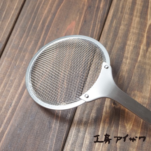 不鏽鋼萬用撈油勺，帶有細緻網孔的圓形濾網，連接至一體成形的不鏽鋼手柄，整體呈現銀色金屬光澤，放置於深棕色木紋背景上。