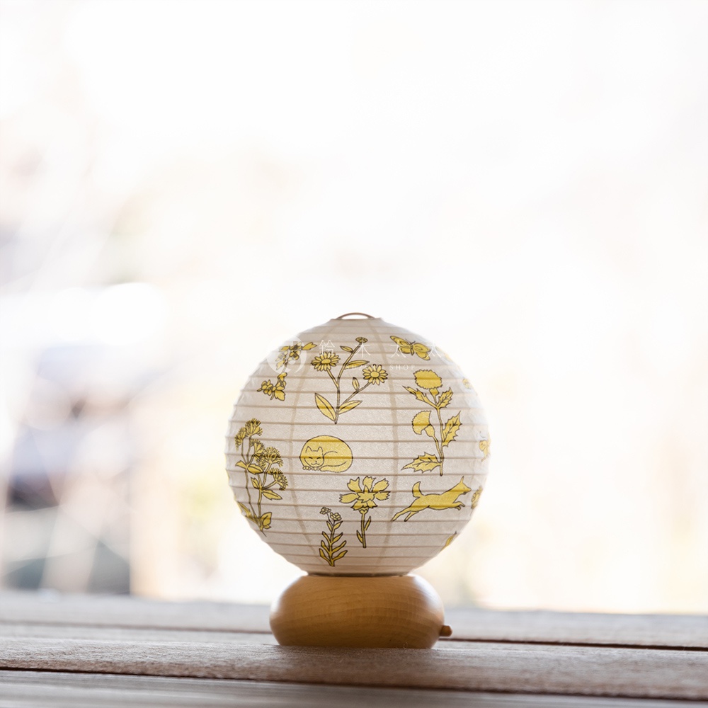 一個白色的紙製圓形提燈，上面印有黃色的花卉、蝴蝶、貓咪和奔跑的狗圖案，提燈底部有一個圓形的木製底座。