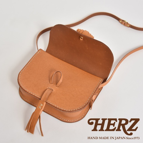 淺棕色皮革流蘇側背包，帶有可調節肩帶和翻蓋設計，側面有流蘇裝飾，包蓋中央有皮革繫帶，整體呈現簡約復古風格，高品質手工製作。