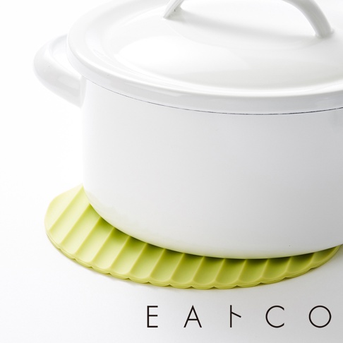 一個淺綠色的圓形隔熱墊，表面有放射狀的溝槽紋理，墊在一個白色金屬鍋的底部，鍋蓋已蓋上，鍋子放在白色背景上。