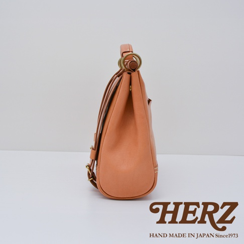 HERZ 職人手作皮革屋瓦風側背包，以溫潤的橘棕色皮革製成，從側面展現包款的立體弧線與精緻縫線細節，下方並有品牌標誌與「HAND MADE IN JAPAN Since1973」字樣。