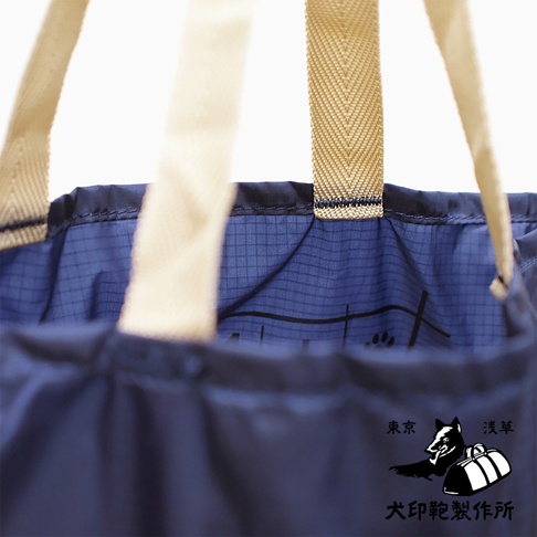 深藍色環保折疊收納購物袋，袋身採用光滑防水尼龍材質，內裡印有網格紋路及狗爪圖案。袋口邊緣縫有黑色滾邊，兩側有米白色帆布提把，提把織帶上有細緻的紋路。