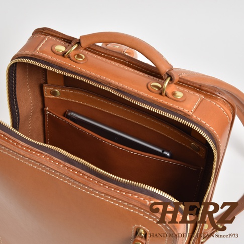 職人手作皮革商務電腦後背包 R-25，採用棕色皮革材質，配有金色拉鍊和金屬扣環。內部有一個手機隔層，手機置於其中。