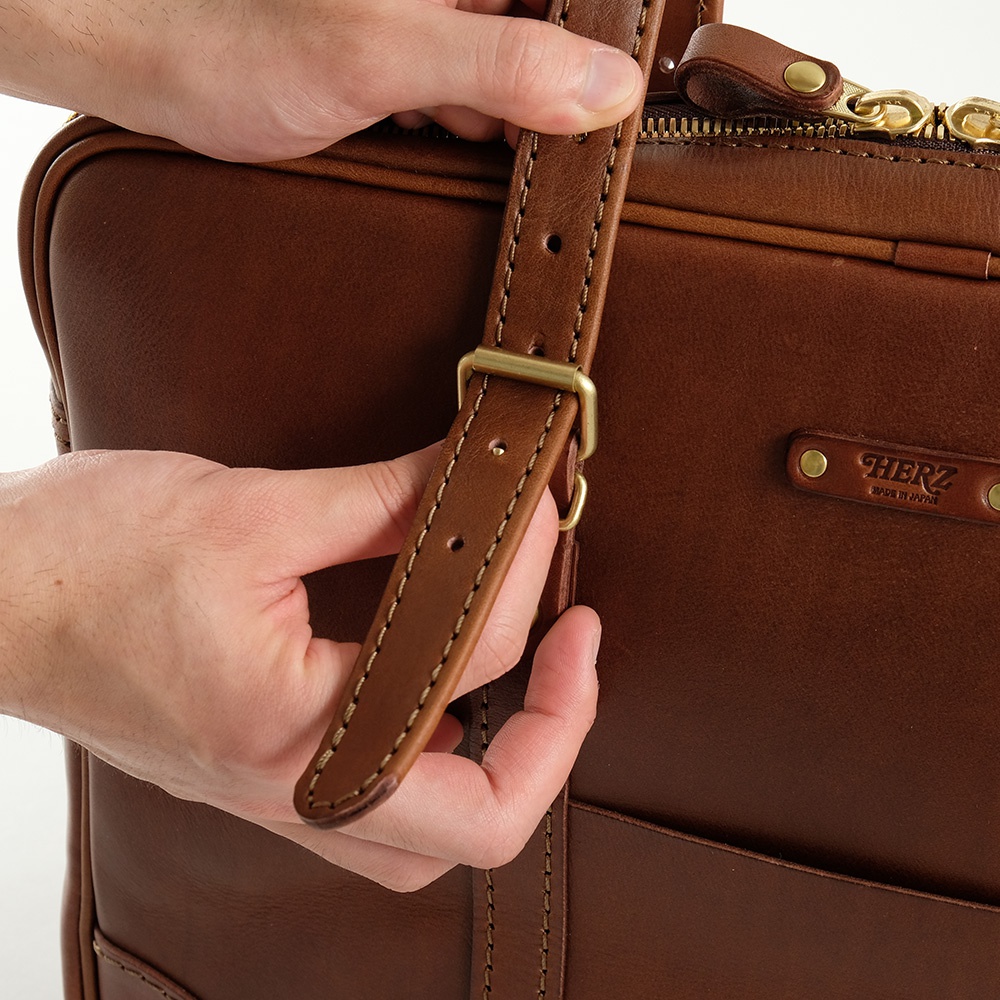 職人手作皮革公事包，棕色皮革，可調節式肩帶，配有金色調節環和扣環。包身有縫線細節，左側有「HERZ MADE IN JAPAN」壓紋標誌。