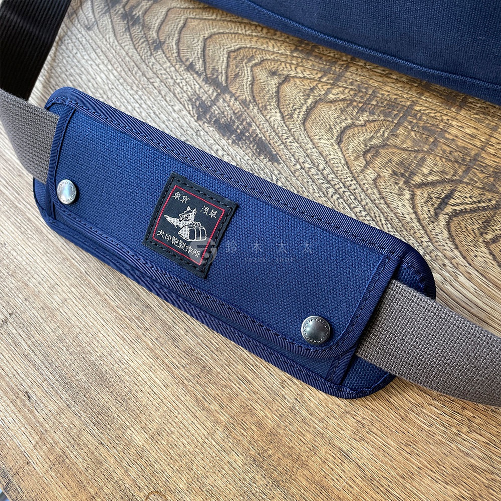 深藍色純棉帆布製成的肩墊，帶有黑色織帶和銀色鉚釘。肩墊中央縫有一個印有「黑犬印」字樣和圖案的紅色標籤。