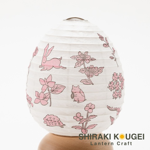 一個白色的紙質提燈，上面印有粉紅色的兔子、花朵和刺蝟圖案，提燈頂部有金屬掛環，下方由木製底座支撐。