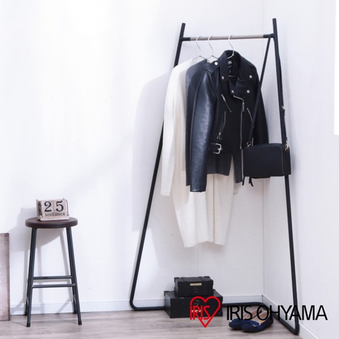 角落造型吊掛衣桿，黑色金屬框架，上方掛有白色長袖毛衣、灰色針織衫和黑色皮夾克，右側掛有黑色方形包，下方有兩個黑色收納盒和一雙藍色平底鞋。