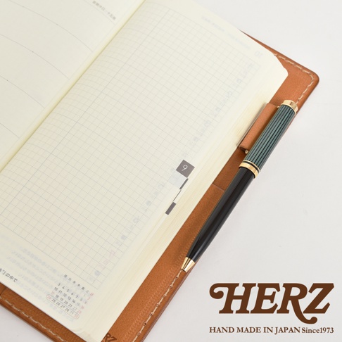 職人手作皮革旅行記事本書套，淺棕色皮革，內含筆記本，筆套中插著一支綠色與黑色筆身的鋼筆。