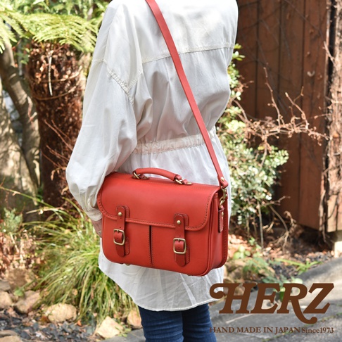 鮮紅色皮革製成的矩形單肩包，帶有頂部提把、可調式斜背帶、雙搭扣前袋和金色金屬配件。