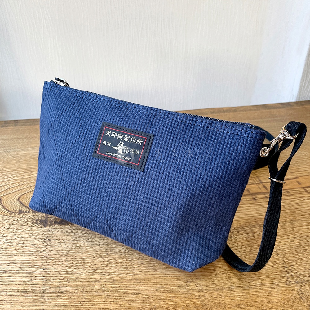 深藍色燈芯絨材質的刺子織斜背包，正面有黑色標籤，印有日文及英文品牌名稱。包包有拉鍊開合，配備黑色織帶和金屬掛勾。